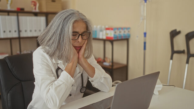 Woman doctor wearing white coat and stethoscope, hands on temples and glasses, seated at desk with laptop in clinic building; work stress.