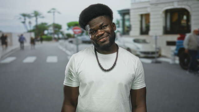 Man smiling with visible face and beaded necklace on a city street crosswalk with blurred pedestrians and cars; confidence charisma ease.