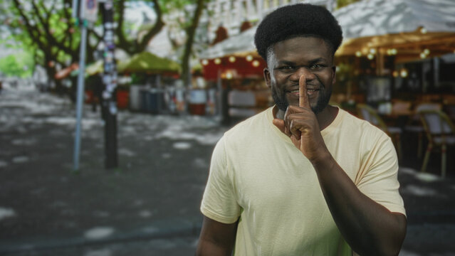 Young man smiling points finger and finger to lips for silence while standing near outdoor cafe on a city street; playful secrecy.