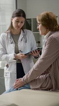 Vertical shot of elderly female patient describing knee pain to young adult physician with tablet during appointment at primary care office