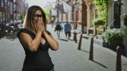 Fototapeta premium Woman covering mouth with hands on street, wearing black blouse and glasses near bollards and storefronts; shock vulnerability.