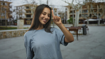 Fototapeta premium Young hispanic woman smiling points finger up on a city street plaza near benches and trees; confidence.