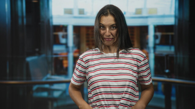 Young hispanic woman grimacing with contorted face and hands at waist inside cruise ship building, appearing queasy and uncomfortable; seasickness disgust.