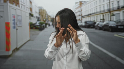 Fototapeta premium Woman with hands raised shielding face on city street wearing white shirt and long hair; distress vulnerability.