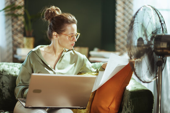 A focused woman with glasses reviews documents while working on her laptop from a green velvet sofa. The running standing fan highlights a productive home office environment in warm weather.