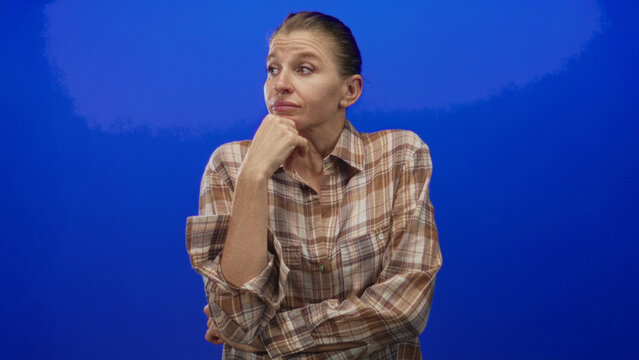 Woman wearing plaid shirt with hand on chin and folded arm, looking up in blue studio set framed from waist; pensive thought curiosity.