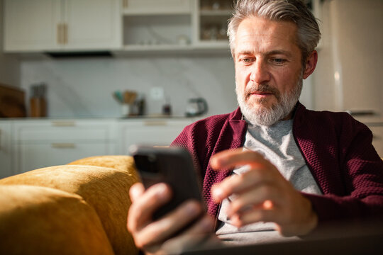 Mature man using smartphone on sofa at home kitchen