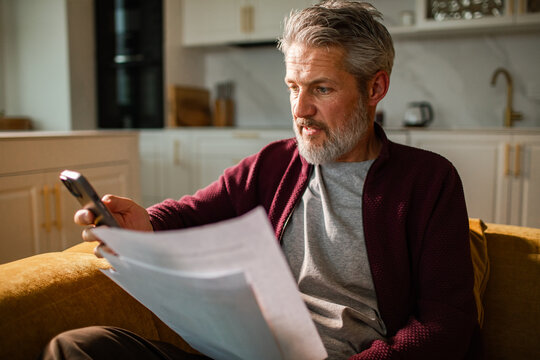 Mature man reviewing bills with smartphone at home kitchen