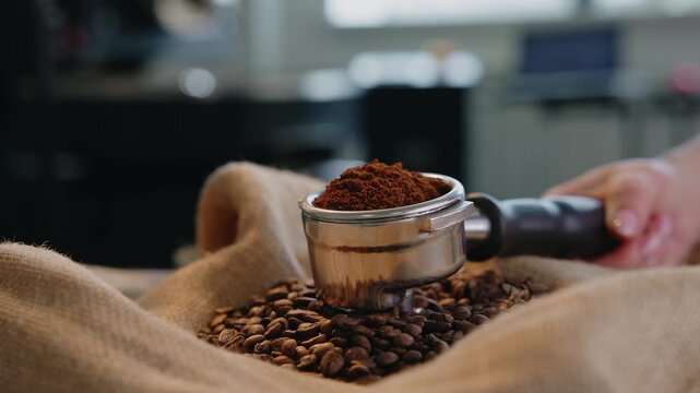Tracking shot portafilter filled with ground coffee placed on burlap sack of roasted beans on cafe countertop with shallow focus.