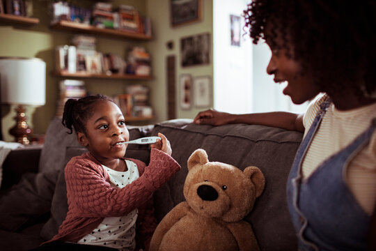 Parent helping sick child take temperature on living room sofa