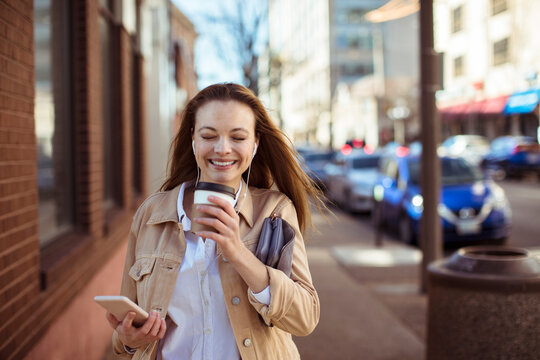Smiling young woman with coffee and smartphone walking on city street