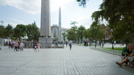 Obraz premium Historic plaza defocused soft bokeh with obelisk, trees and broad paved walkway in shallow focus; backdrop copyspace calm.
