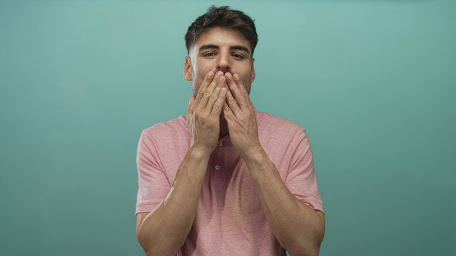 Man in pink polo shirt gently blows a kiss with hand extended in a turquoise studio setting; affection.