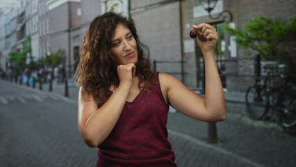 Fototapeta premium Woman holding cherry between fingers with chin on fist on street, raising arm and smiling while wearing burgundy sleeveless top; playful curiosity.