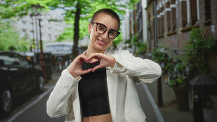 Fototapeta premium Woman with short hair and glasses forms a heart with hands over chest wearing white blazer on an urban street; affection.