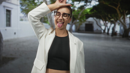 Fototapeta premium Woman in glasses and white blazer sticks out tongue and raises hand to forehead on sunlit street with trees; playful carefree attitude.