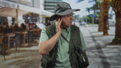 Obraz premium Man in fishing vest and wide hat, hand to cheek while grimacing and closing eyes on a palm lined street beside outdoor cafe; discomfort.