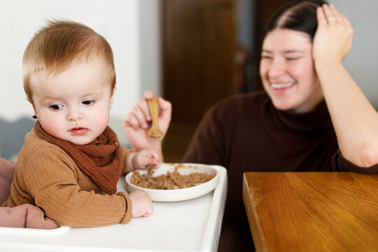 Happy Mother feeding cute baby infant in high chair with complementary food. Family domestic life. Mom and son having dinner together, joyful moment. Happy Mother's day!