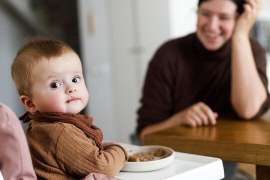 Happy Mother feeding cute baby infant in high chair with complementary food. Family domestic life. Mom and son having dinner together, joyful moment. Happy Mother's day!
