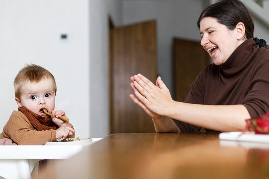 Happy Mother feeding cute baby infant in high chair with complementary food. Family domestic life. Mom and son having dinner together, joyful moment. Happy Mother's day!