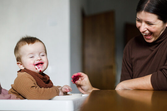 Happy Mother feeding cute baby infant with fuit in high chair, eating complementary food. Family domestic life. Mom and son together, joyful moment. Happy Mother's day!