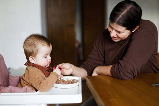 Mother feeding cute baby infant with fuit in high chair, eating complementary food. Family domestic life. Mom and son together, joyful moment. Happy Mother's day!