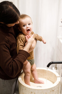 Mother cleaning cute baby infant in modern bathroom after eating complementary food. Family domestic life. Mom washing son face with water in sink, joyful moment. Happy Mother's day!