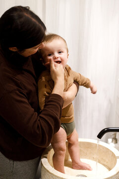 Mother cleaning happy baby infant in modern bathroom after eating complementary food. Family domestic life. Mom washing son cute face with water in sink, joyful moment. Happy Mother's day!