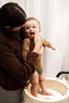 Mother cleaning happy baby infant in modern bathroom after eating complementary food. Family domestic life. Mom washing son cute face with water in sink, joyful moment. Happy Mother's day!