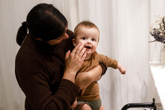 Mother cleaning happy baby infant in modern bathroom after eating complementary food. Family domestic life. Mom washing son cute face with water in sink, joyful moment. Happy Mother's day!
