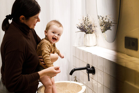 Mother cleaning happy baby infant in modern bathroom after eating complementary food. Family domestic life. Mom washing son cute face with water in sink, joyful moment. Happy Mother's day!