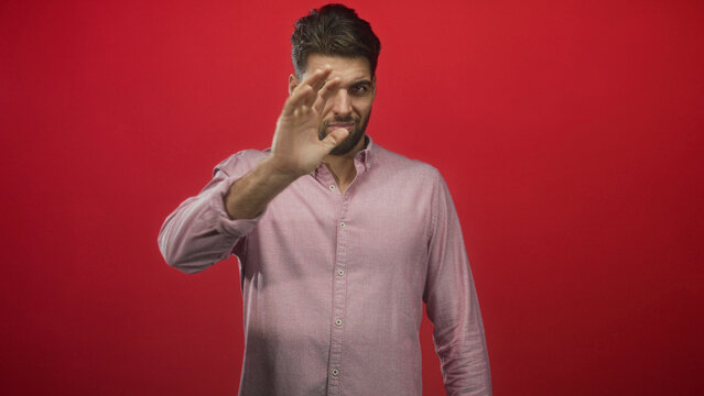 Hispanic man with beard covering face with hand in studio against red backdrop; embarrassment privacy.
