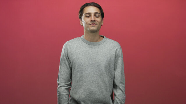 Young man shrugs shoulders with eyes closed wearing a gray sweater, hands near pockets in a studio; calm nonchalance.