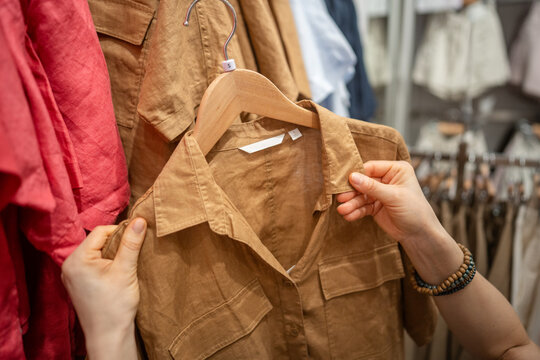 Woman customer shopping for clothes in fashion shop, choosing outfit from new collection, female client holding hanger with shirt checking item quality, content of fabric and size in apparel showroom