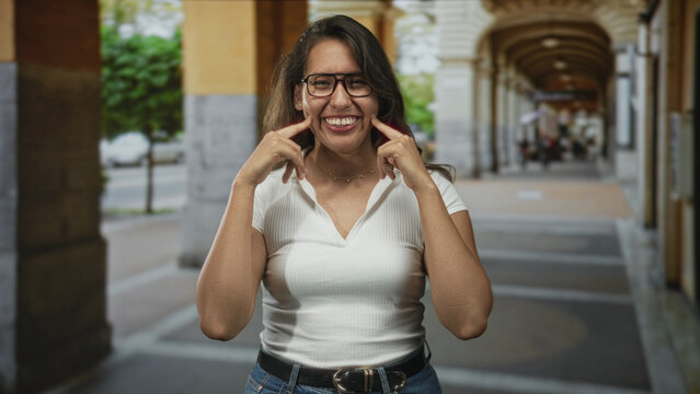 Woman points fingers to cheeks smiling with glasses and bare forearms in a covered street arcade with stone arches and blurred pedestrians; joy simplicity.