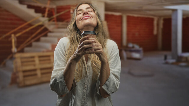 Middle age woman holding coffee cup with hands and closed eyes in unfinished building renovation site; calm reflection.