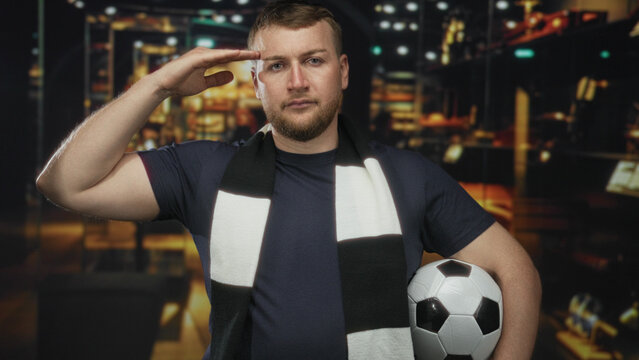 Man salutes hand to forehead holding a soccer ball in a shop building with black and white scarf; pride fandom loyalty.