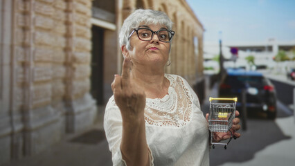 Obraz premium Woman in white lace blouse wearing glasses holding a miniature metal shopping cart with clenched hand on street by stone building and parked car; defiance.