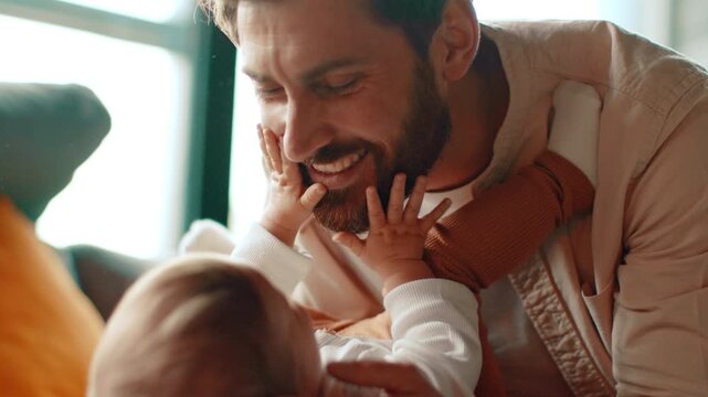 A man with a beard is having fun with his baby, lifting them gently while they both smile. The living room has large windows letting in light, creating a warm atmosphere.