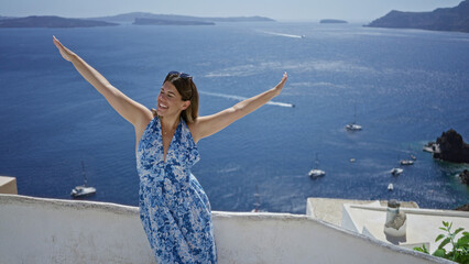 Obraz premium Woman smiling with arms out on building terrace overlooking sea and boats in santorini town; vacation relaxation joy.