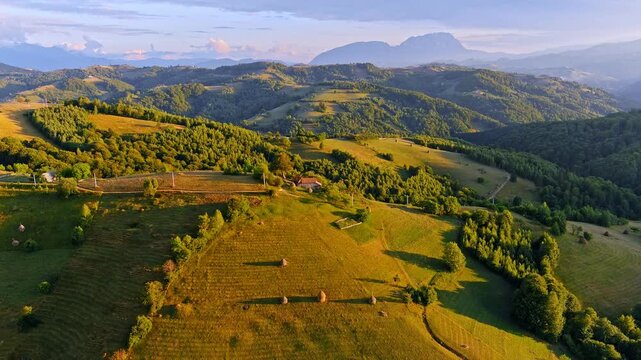 Aerial view of the rolling hills and haystacks of Transylvania with scattered houses and dense forests during golden hour in Holbav, Brasov, Romania.