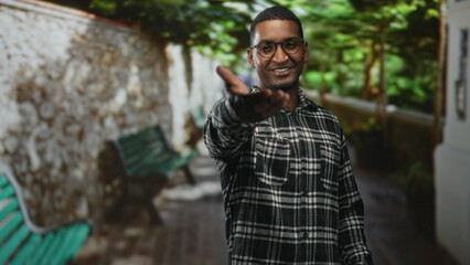 Fototapeta premium Young black man reaches out with open hand toward camera on a leafy street, smiling and wearing glasses and plaid shirt; friendly welcome.