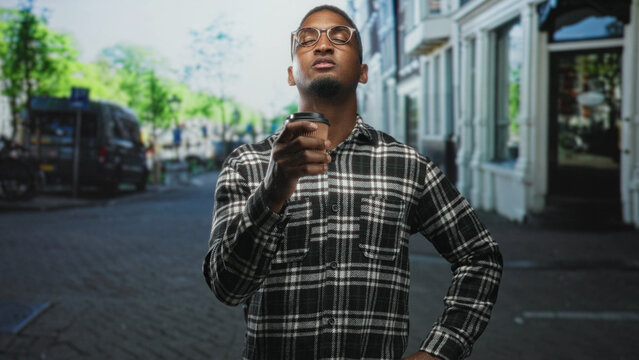 Young black man holding coffee cup near face with eyes closed, wearing flannel shirt and glasses, standing on street in front of a storefront; quiet contemplation.