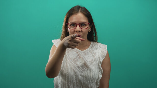 Woman with red glasses pointing finger in studio against teal backdrop wearing white lace blouse, direct gesture; assertive challenge.