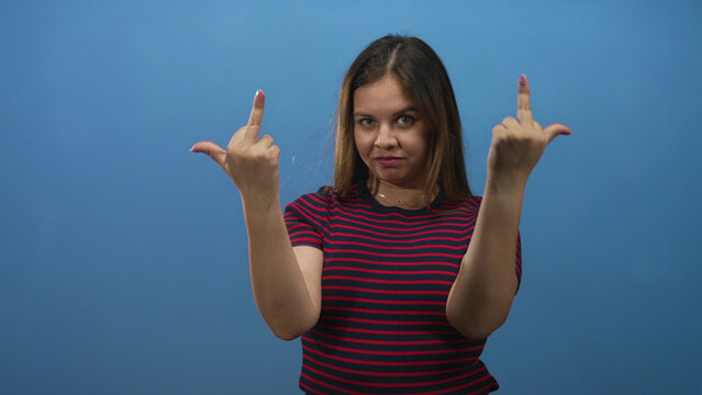 Young woman shows middle finger and raised hand toward camera in blue studio setting; defiance bold attitude.