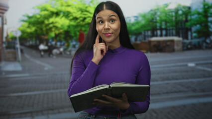 Fototapeta premium Young latina woman in purple top holds hardcover book and hand on chin gesture on street; curiosity.
