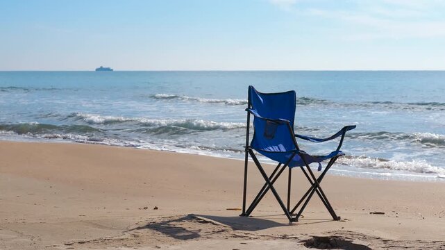 Solitary blue camping chair sitting on a sandy beach with gentle waves lapping the shore on a sunny day. A ship sailing on the distant horizon adds to the tranquil and relaxing atmosphere