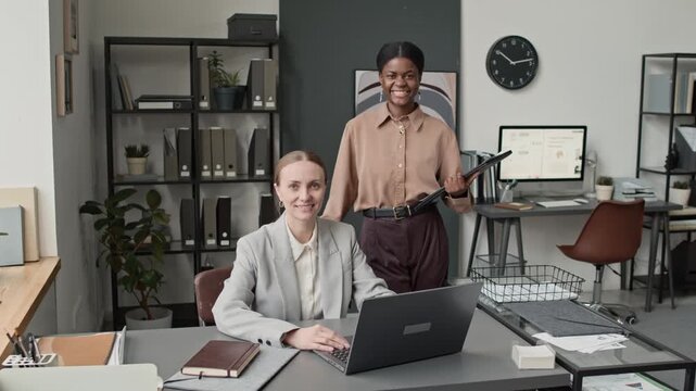Portrait shot of Caucasian woman sitting at her laptop, and African American woman standing next to her with folder in modern business office