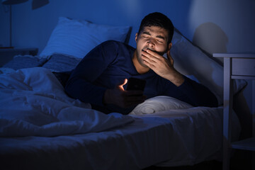 Fototapeta premium Sleepy young man using mobile phone in bed at night
