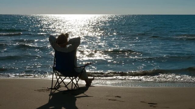 Enjoy the sunset on the beach. Silhouetted woman sitting on a camping chair, relaxing with hands behind her head while enjoying the beautiful sunset view over the glittering sea and gentle waves
